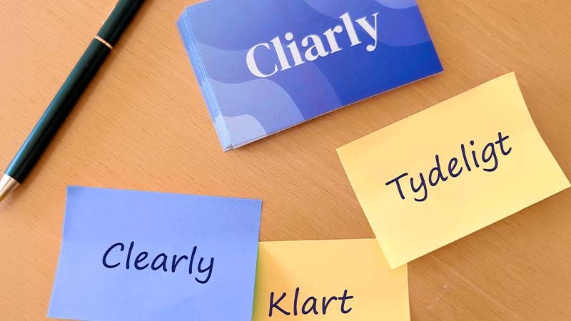 Et skrivebord med en kuglepen, Cliarly-visitkort og tre post-its med ordene 'Clearly', 'Tydeligt' og 'Klart', som illustrerer betydningen af virksomhedens navn.