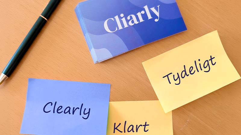 Et skrivebord med en kuglepen, Cliarly-visitkort og tre post-its med ordene 'Clearly', 'Tydeligt' og 'Klart', som illustrerer betydningen af virksomhedens navn.
